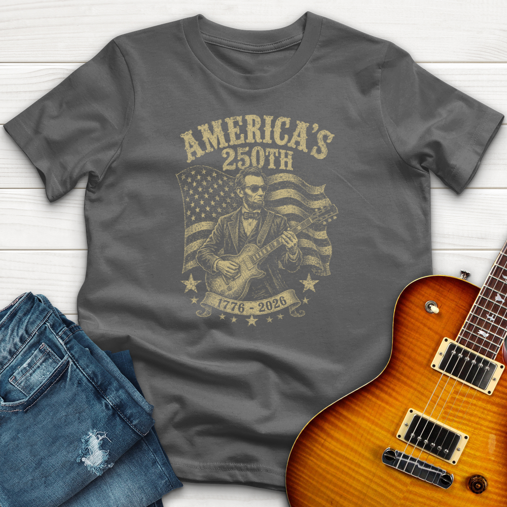 Gray t-shirt with graphic design of a person playing guitar and American flag, next to jeans and a guitar on a wooden surface.