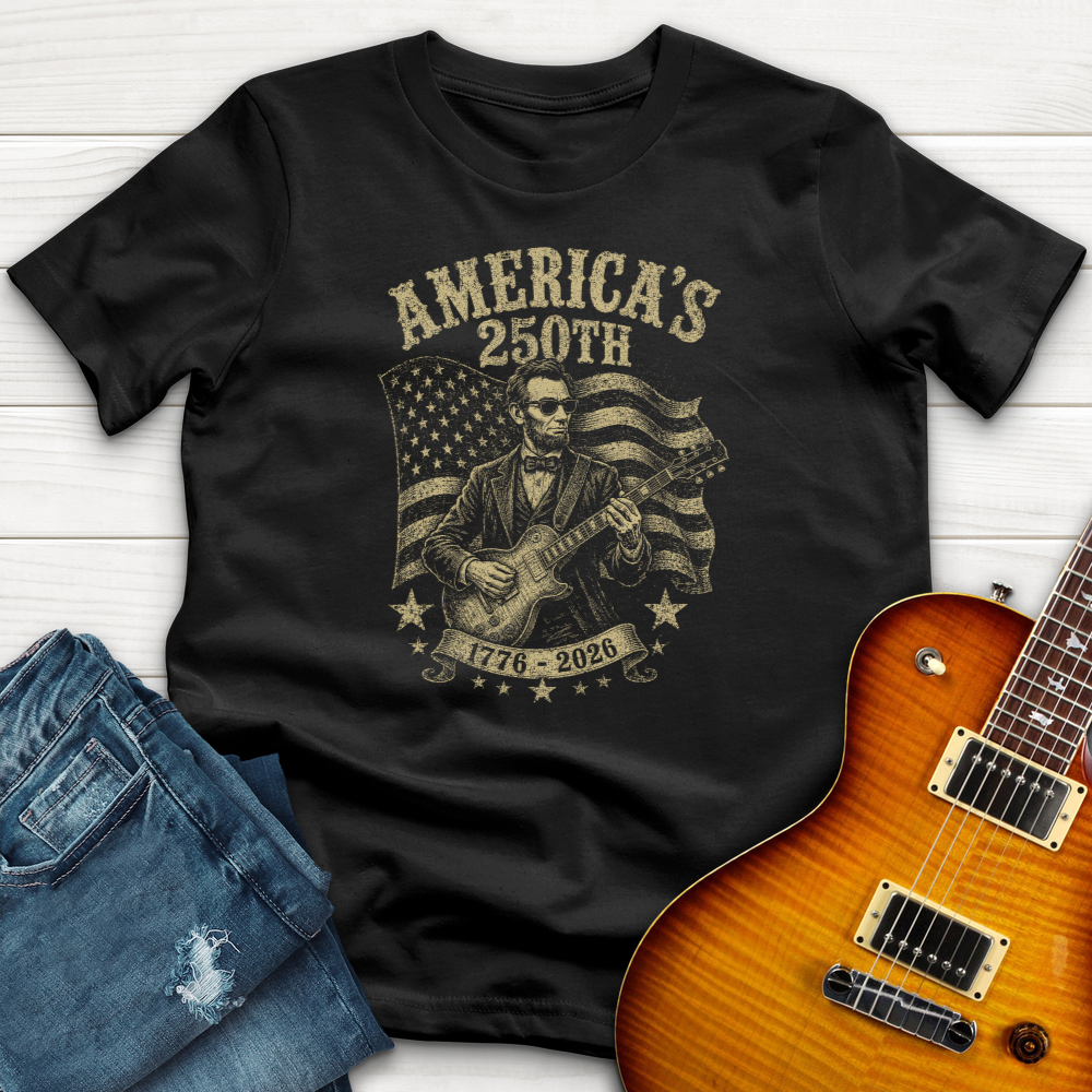 Black t-shirt with graphic design of a person playing guitar and American flag, placed on a wooden surface with a guitar and jeans beside it.