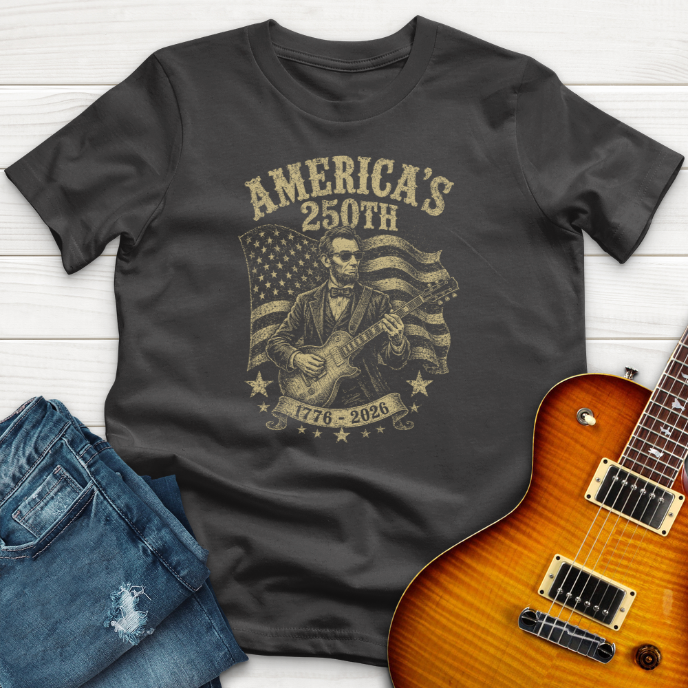 Gray t-shirt with graphic design of a person playing guitar and American flag, next to jeans and a guitar on a white wooden surface.